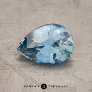 A pear-shaped blue gemstone (Product Code: JM1028L) rests on a textured surface. The image includes the logo and the text "Earth's Treasury" at the bottom.