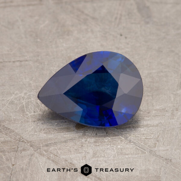 A polished, pear-shaped blue gemstone rests on a textured surface with the product name "JM1034L" at the bottom.