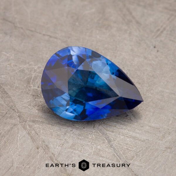 Pear-shaped blue sapphire gemstone (JM1035L) resting on a textured gray surface with the "Earth's Treasury" logo at the bottom.