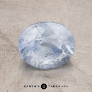 A 2.98-carat, oval-cut light blue sapphire with visible inclusions rests on a textured beige surface. The phrase "Earth's Treasury" appears at the bottom.