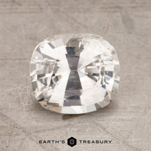 A 2.23-carat, cushion-cut white sapphire rests elegantly on a textured gray surface, adorned with the "Earth's Treasury" logo at the bottom.