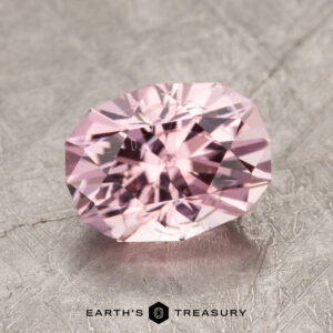 An ET1614 gemstone with a faceted pink oval cut resting on a textured metallic surface bears the label "EARTH'S TREASURY.