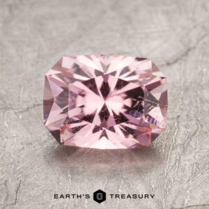 A rectangular pink gemstone with brilliant facets placed on a textured grey surface. Text at the bottom reads "ET1625 Earth's Treasury.