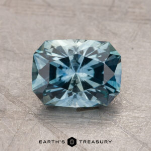 A 2.14-carat Montana Sapphire (heated), featuring a rectangular, faceted blue gemstone, is placed on a textured surface. The bottom center displays the logo "Earth's Treasury.