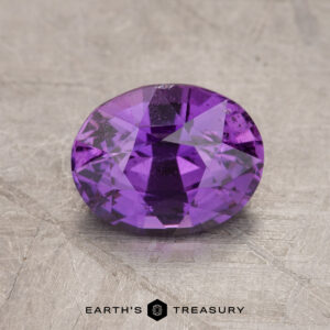 A polished, oval-shaped 2.01-carat sapphire gemstone with a deep purple hue, set on a textured grey surface. The name "Earth's Treasury" is visible below the gemstone.