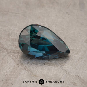 Close-up of a dark blue to green faceted 1.55-carat sapphire in a pear shape, placed on a textured gray surface. Text below reads "EARTH'S TREASURY" with a small hexagon symbol.