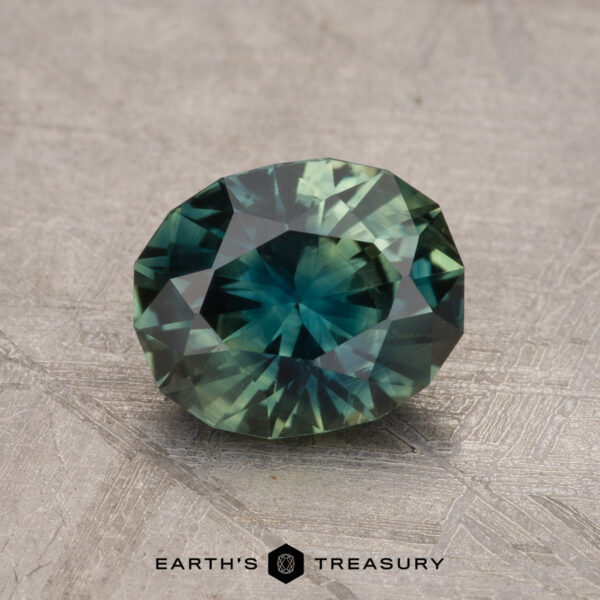 A 1.61-carat heated Australian sapphire, featuring a faceted oval shape with shades of green and blue, is displayed on a textured surface with the text "Earth's Treasury" at the bottom.