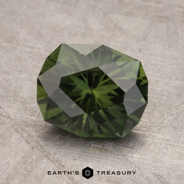 A close-up of a faceted green gemstone resting on a textured surface with the "Earth's Treasury" logo at the bottom, identified as a 2.20-Carat Australian Sapphire (Heated).