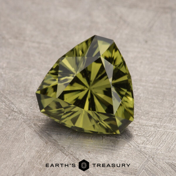 A 1.77-carat Australian sapphire, heated and faceted in a triangular shape, is placed on a textured metallic surface. The gem is labeled "Earth's Treasury.