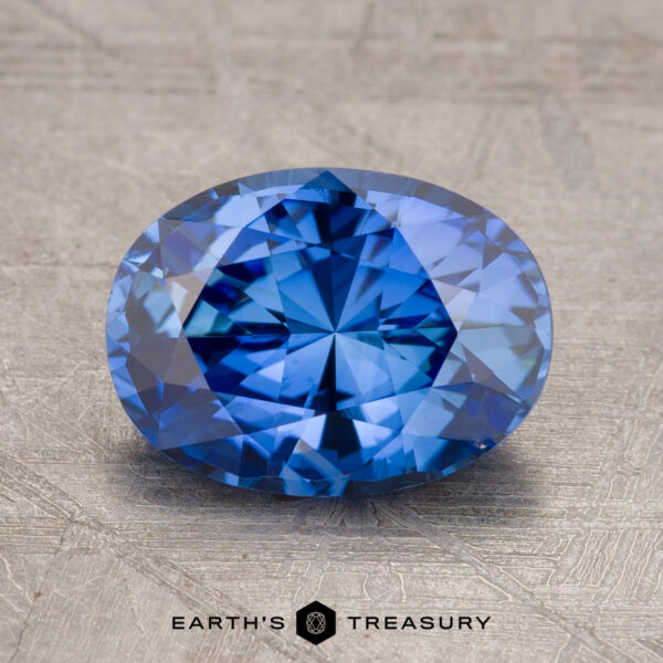 A faceted blue gemstone, the 2.47-Carat Madagascar Sapphire, oval-shaped and displayed on a textured grey surface with the logo "Earth's Treasury" below it.