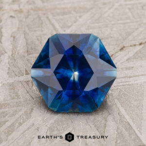 A faceted, six-sided blue gemstone placed on a textured surface. The text "EARTH'S TREASURY" is displayed below the 2.06-Carat Royal Blue Montana Sapphire (Heated).