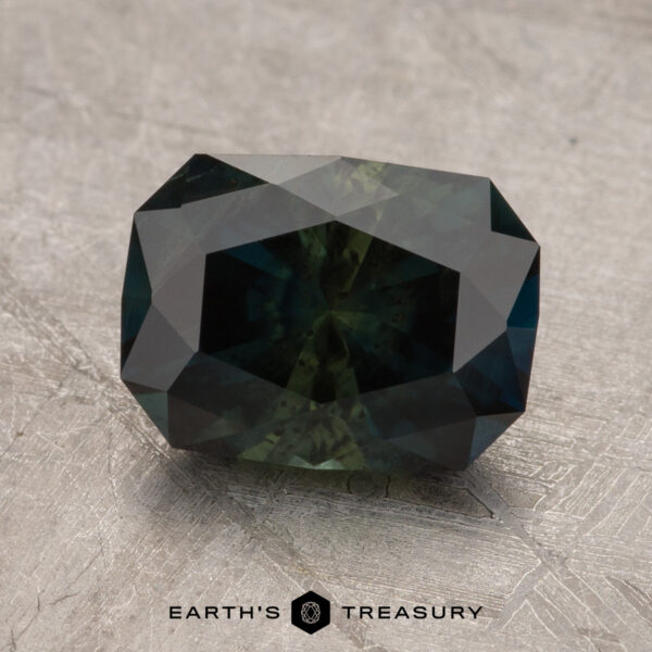 A rectangular-cut, dark-hued Australian Sapphire weighing 2.55 carats rests on a textured surface, with the text "EARTH'S TREASURY" displayed at the bottom.