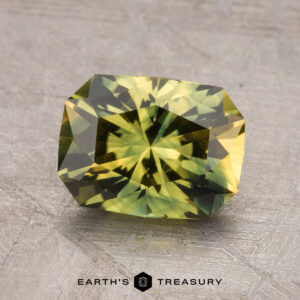 A 2.24-Carat Australian Sapphire, with green facets and a yellow tint, sits on a textured gray surface. The text "EARTH'S TREASURY" is visible below the stone.