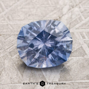 A 2.53-carat Violet-Blue Umba Sapphire, with intricate faceting, is placed against a textured light grey background, with the text "EARTH'S TREASURY" at the bottom center.