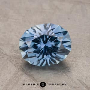 A faceted 2.20-carat Montana Sapphire, radiating a medium blue hue, is positioned on a textured surface. Below the gemstone, the text "Earth's Treasury" is visible.