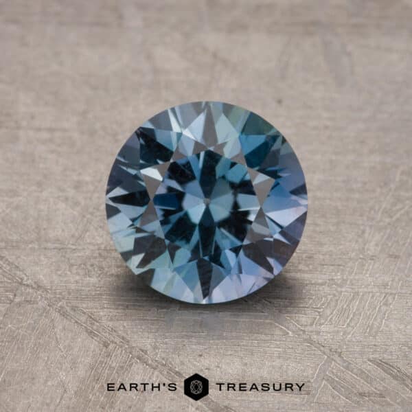 A round-cut, 1.73-carat Montana sapphire boasting a captivating blue and white color gradient is displayed on a beige textured surface. The text "Earth's Treasury" is featured at the bottom.