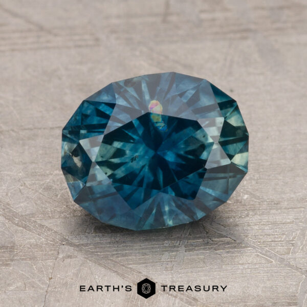 A faceted dark blue gemstone, specifically a 2.46-carat Montana Sapphire (Heated), is placed on a textured surface. The text "EARTH'S TREASURY" is visible beneath the stone.