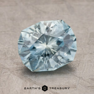 A faceted, light blue 2.43-carat Montana Sapphire (heated) is placed on a textured gray surface. The words "Earth's Treasury" are displayed at the bottom.