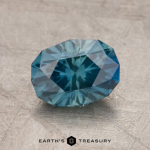 A faceted 2.00-carat Montana Sapphire (heated) gemstone on a textured gray surface with "Earth's Treasury" text at the bottom.