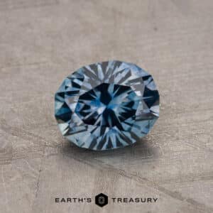 A round cut 1.21-carat Montana Sapphire (Heated) displayed on a textured surface with the text “EARTH’S TREASURY” at the bottom.