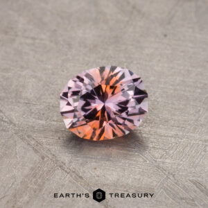 A 0.65-carat Montana sapphire (heated) showcasing a mix of pink and orange hues, resting on a textured surface with the text "Earth's Treasury" at the bottom.