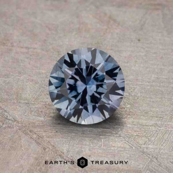 A 0.84-carat Montana Sapphire (Heated) with intricate facets is displayed on a textured surface, showcasing its steely blue hue. The text "EARTH'S TREASURY" is visible at the bottom.