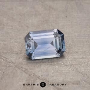 A stunning 0.61-Carat Montana Sapphire (Heated), emerald-cut and delicately placed on a textured gray surface. Text at the bottom reads "EARTH'S TREASURY.