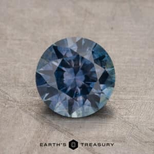 A round, 1.54-carat heated Montana Sapphire rests on a textured surface with the logo and text "Earth's Treasury" below it.