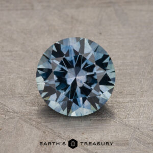 A round, faceted 1.60-carat Montana Sapphire (Heated) with shades of blue sits on a textured surface, with "Earth's Treasury" written at the bottom in black text.