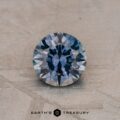 A round faceted gemstone with a steely blue and gray hue, resting on a textured surface. The text "Earth's Treasury" is displayed below the 0.92-Carat Montana Sapphire (Heated).