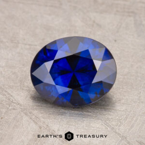 A round, faceted 1.87-carat sapphire on a textured gray surface with the label "Earth's Treasury" at the bottom.