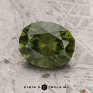 A 2.51-carat olive green Australian sapphire (heated) is displayed on a textured gray surface. The image features the logo and text "EARTH'S TREASURY" at the bottom.