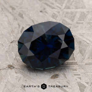 A 2.29-carat Midnight Blue Australian Sapphire (Heated), displayed on a textured gray surface, with the text "EARTH'S TREASURY" below.