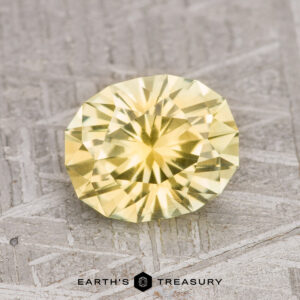 A round, faceted 2.13-carat light yellow Montana sapphire (heated) is placed on a textured grey surface. The text "EARTH'S TREASURY" is written below the gemstone.