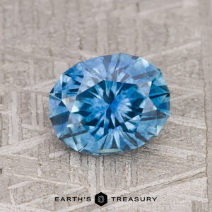 A 1.86-carat, medium blue Montana sapphire with intricate faceting is displayed on a textured gray surface with the text "EARTH'S TREASURY" at the bottom.