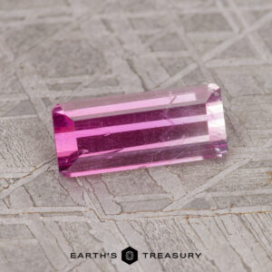 A 1.24-carat hot pink-pale pink bicolored Umba sapphire, with a rectangular, faceted shape, lies on a grey textured surface. The text "EARTH'S TREASURY" appears at the bottom of the image.