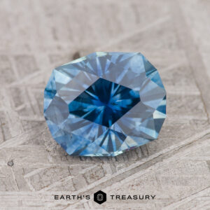 A faceted teal blue Montana sapphire, weighing 2.10 carats and displayed on a textured surface, with the text "EARTH'S TREASURY" visible at the bottom.