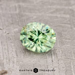 A 0.84-carat mint green demantoid garnet with a round, faceted design is showcased on a textured gray surface. Below it, you'll find the Earth's Treasury logo.