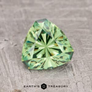 A faceted, triangular 1.57-carat medium green demantoid garnet rests on a textured gray surface. Text reads: "Earth's Treasury.