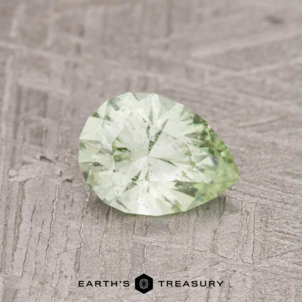 The 0.80-carat pale kiwi green grossular garnet, featuring a faceted pear shape, rests on a textured surface.