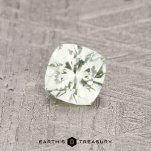 A cushion-cut pale kiwi-green gemstone, weighing 0.47 carats, resting on a textured gray surface with the "Earth's Treasury" logo at the bottom.