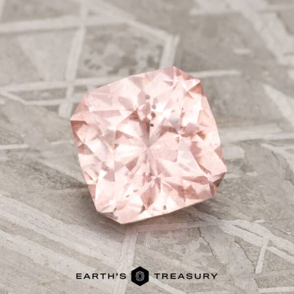 A 1.84-carat pink-peach pastel pyrope garnet is showcased on a textured gray surface, with the text "Earth's Treasury" at the bottom.