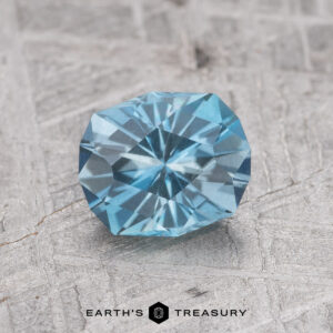 The 0.92-Carat Nasarawa Teal Aqua, a round, faceted blue gemstone, is showcased on a textured gray background with the Earth’s Treasury logo visible at the bottom.