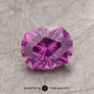 A faceted ET1123 gemstone in purple rests on a textured gray surface.