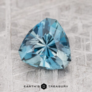 A 1.50-carat Nasarawa Teal Aqua gemstone, with intricate facets and a triangular cut, is displayed on a textured surface.