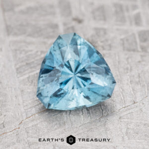 A trillion-cut 0.99-carat Nasarawa Teal Aqua gemstone on a textured gray surface, featuring the "Earth's Treasury" logo at the bottom.