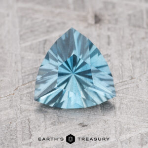 A 0.95-carat Nasarawa teal aqua gemstone, with intricate triangular cuts, rests on a textured gray surface. Text at the bottom reads "Earth's Treasury.