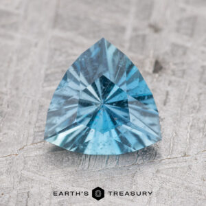 A 1.11-carat Nasarawa teal aqua trillion-cut gemstone with visible facets, displayed on a textured surface.