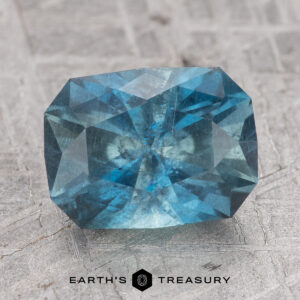 The 2.63-Carat Nasarawa Teal Aqua sapphire, with its rectangular shape and slightly rounded corners, is showcased on a textured gray surface.