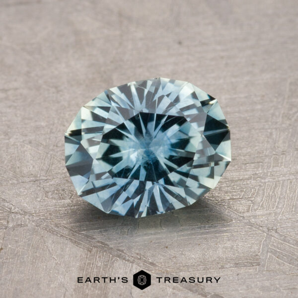 A 1.47-carat Montana Sapphire, round and faceted in light blue with an intricate pattern, is elegantly displayed on a textured gray surface.